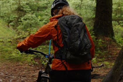 female adult volunteer walking sitting on a mountain bike in the forest wearing orange jacket with black backpack and black helmet