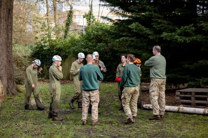 group of cadets and volunteers stood in a circle wearing green overalls and white helmets in a field next to the woods