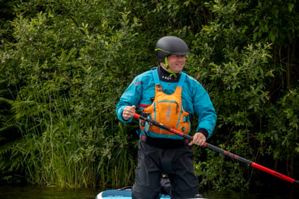 Adult volunteer on their knees on a paddle board floating across a lake wearing a blue jacket and yellow buoyancy aid and black helmet.