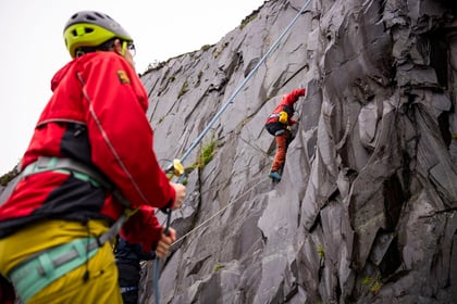 Person climbing a ruggid cliff being supported by someone stood on the ground wearing a red jacket and yellow helmet