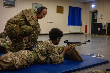male cadet in military uniform on a blue mat with a supported air rifle. An adult in military uniform kneeling next. to the cadet giving direction.