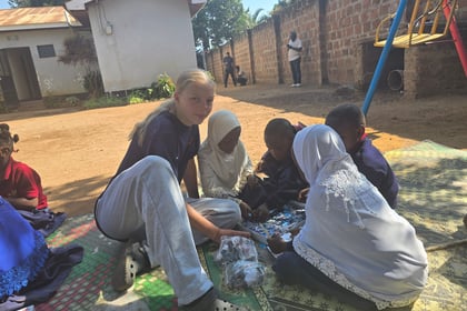 Female cadet sits on grass mats while playing with a puzzle with kids from a local orphanage