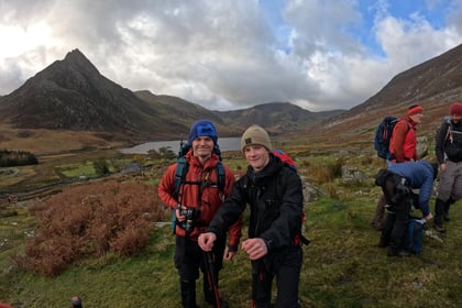 Two male cadets smile for a photo with mountains in the distance wearing walking gear