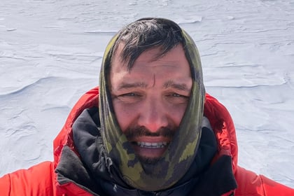 Jordan Wylie stands on glacier with mountain peaks behind him wearing an orange jacket