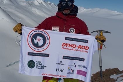 Jordan Wylie holds flag with various sponsor logos on it at the top of a mountain summit