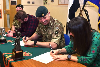 3 people sat at desk signing paper, middle person in military uniform smiling