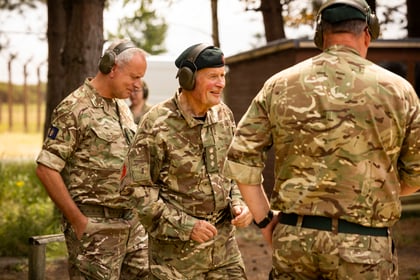 Three men in camouflage military uniforms and ear protection stand and converse outdoors near trees and a building on a sunny day.