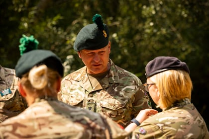 A group of Army Cadet personnel in camouflage uniforms and green berets stand outdoors, engaged in conversation. Sunlight filters through the trees, casting light on their faces.