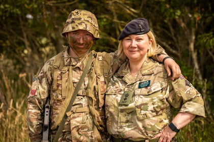 Two Army Cadets in camouflage military uniforms stand outdoors, one with face paint and a helmet, the other with a beret. They are smiling, with the person on the left’s arm round the other’s shoulder. Trees and grass are in the background.