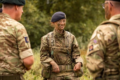 A group of Army Cadets in camouflage uniforms and berets stand outdoors, one woman in the centre with face paint, smiling and talking to others, with greenery in the background.