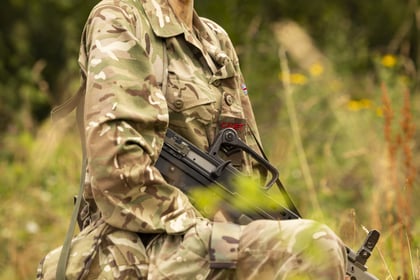 A person in camouflage Army Cadet uniform and hat, with face paint, kneels and smiles outdoors amid tall grass and greenery, holding a rifle.