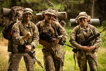 Three smiling Army Cadets in camouflage uniforms and face paint stand outdoors in a forest, holding rifles and carrying large rucksacks with rolled sleeping mats.