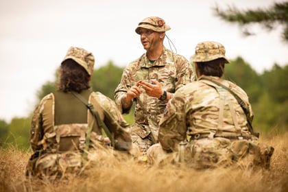 Three Army Cadets in camouflage uniforms sit in dry grass, listening to another soldier who is speaking and gesturing with his hands during a field training session, with a forest in the background.
