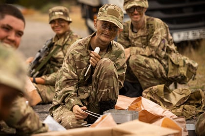 A group of Army Cadets in camouflage uniforms sit on the ground outdoors, smiling and eating with cutlery. One person in the centre holds a fork and looks at the camera, while others sit and relax nearby.