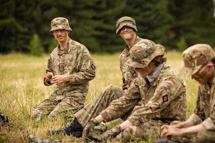 Four Army Cadets in camouflage uniforms and hats sit on grass in a field, smiling and preparing kit, with trees blurred in the background. Their faces are painted with camouflage.
