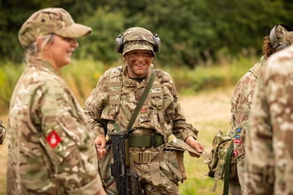 Army Cadets in camouflage uniforms stand outdoors. One in the centre smiles with a rifle and face paint. Others, slightly out of focus, talk nearby. The background is grassy with trees.