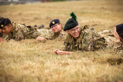 Four Army Cadets in camouflage uniforms lie on dry grass, smiling and chatting. One person in the foreground wears glasses and a green beret with a feather. The others wear dark berets. All appear relaxed and happy.