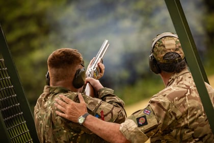 Two people in camouflage uniforms and ear protection, one guiding the other who is aiming a shotgun outdoors, with green foliage in the background.