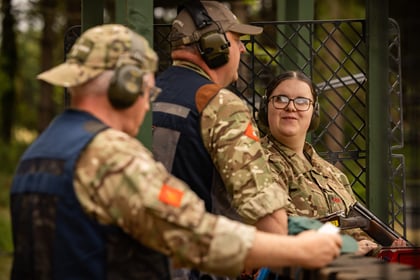 Three people in camouflage and ear protection, talking at an outdoor shooting range. One woman holds a rifle and smiles at a man, whilst another man stands nearby, all engaged in conversation.