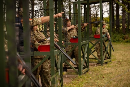 Five Army Cadets in camouflage uniforms participate in a shooting activity outdoors, standing in separate green bays and aiming shotguns at targets. Trees and grass are visible in the background.