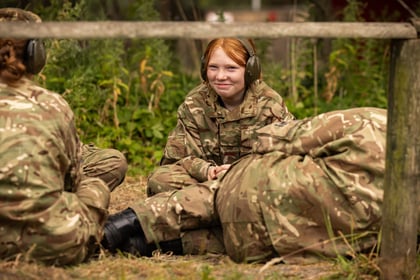 A young person in camouflage uniform and ear defenders sits outdoors, smiling, with others in similar attire nearby. The group is surrounded by greenery and appears to be taking a break or participating in an outdoor activity.