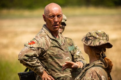 A military officer in camouflage uniform stands outdoors, looking serious, whilst talking to another soldier. Two soldiers are visible, with a blurred field and greenery in the background.