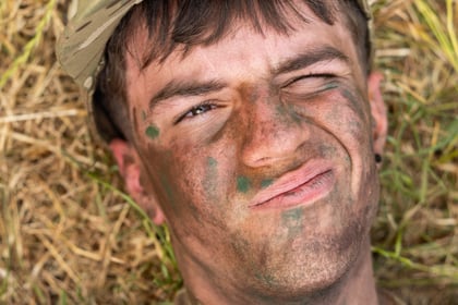 A person in camouflage clothing lies on dry grass, face smeared with dirt, squinting and pulling a face.