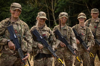 Five Army Cadets in camouflage uniforms stand in a row outdoors, holding rifles with yellow attachments, and have face paint on. Trees and grass are visible in the background.