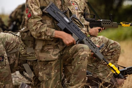 Two Army Cadets in camouflage uniforms and face paint kneel on dry grass, holding rifles with yellow covers on the barrels. Trees and sky are visible in the background.