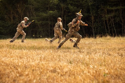 Three Army Cadets in camouflage uniforms run across a dry grassy field with trees in the background, holding rifles as part of a training exercise.