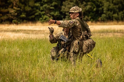 Two Army Cadets in camouflage uniforms crouch in a grassy field. One is pointing forward, while the other looks in the same direction. Trees and a yellow field are visible in the background.