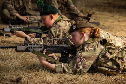 Two young Army Cadets in camouflage uniforms lie on dry grass, aiming rifles during training. One has a green beret with a pom-pom, the other a brown beret. Both have a UK flag patch on their sleeves.
