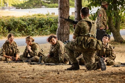 A group of Army Cadets in camouflage uniforms sit on dry grass, listening to a kneeling instructor holding a rifle. Trees and a building are in the background.