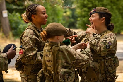 Three young Army Cadets in camouflage uniforms smile and laugh together outdoors, one adjusting another’s jacket while trees and blurred greenery are visible in the background.