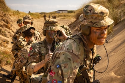 A group of Army Cadets in camouflage uniforms and face paint stand in a sandy, grassy area, some using radios, focussed on their tasks during a military exercise.