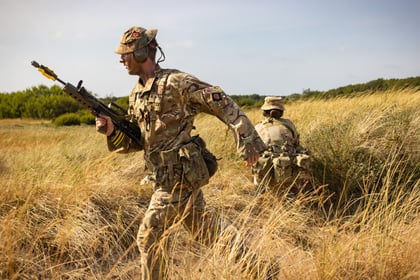 Two Army Cadets in camouflage uniforms move through tall, dry grass. One is running whilst holding a rifle, and the other is crouched down, partially hidden by the grass. Trees and blue sky are visible in the background.