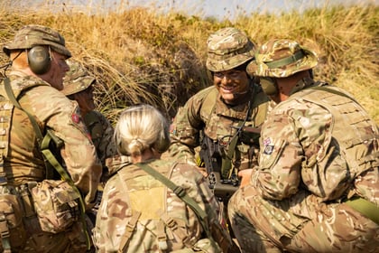 Five Army Cadets in camouflage uniforms and helmets gather outdoors in tall grass, appearing to discuss or plan. One soldier in the centre smiles, while others listen attentively. Sunlight highlights the group and dry vegetation.