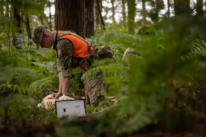 A soldier in camouflage and an orange waistcoat performs CPR on a manikin in a forest, with a laptop displaying monitoring data nearby. Ferns and trees surround the scene.
