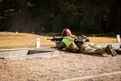 A person in camouflage and a green vest lies prone on a gravel range, aiming a rifle towards targets in a grassy, wooded outdoor setting, with numbered markers visible.