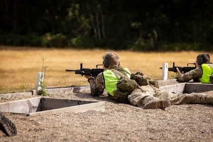 Two people in camouflage uniforms and neon vests lie prone on a gravel field, aiming rifles towards targets. Trees and a grassy area are in the background.