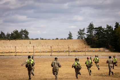 Eight Army Cadets in camouflage uniforms and high-visibility vests run across a dry field towards a shooting range with numbered targets, under a partly cloudy sky. Trees and a large earth berm are in the background.