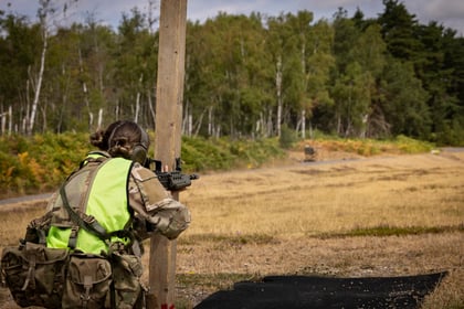 A person in military gear and a neon vest kneels behind a wooden post, aiming a rifle at a distant target in an open, grassy area with trees in the background.
