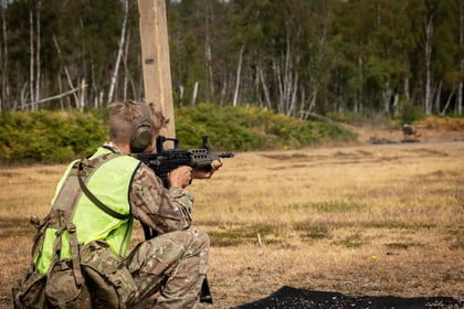 A soldier in camouflage and a neon vest kneels on dry grass, aiming a rifle at a distant target during a training exercise in a wooded outdoor area.