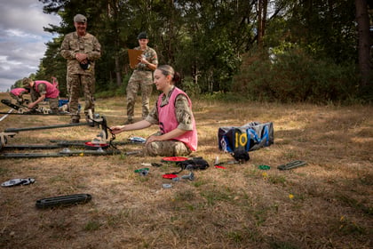 A woman in military uniform and a pink vest kneels on grass, assembling equipment. Two other soldiers, one with a clipboard, supervise nearby. Trees and a blue bag are in the background.