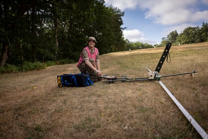 A person in outdoor clothing kneels on a grassy field beside scientific equipment and a blue bag, with trees and a partly cloudy sky in the background.