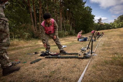 A person in military camouflage and a pink waistcoat operates a small catapult outdoors, launching an object across a grassy field, with others working in the background near trees.