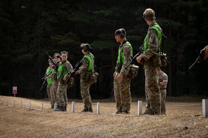 Seven soldiers in camouflage uniforms with green vests and ear protection stand in a line on a grassy field, with trees in the background. Some hold rifles, and all appear to be focused on an unseen target.