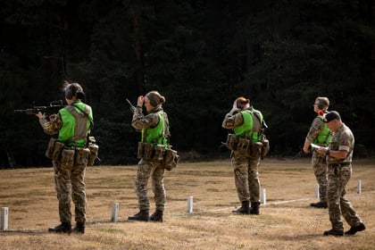 Four people in Army Cadet uniforms and green vests stand on a grassy field; two hold rifles while one uses binoculars and another writes on a clipboard. Trees are in the background.