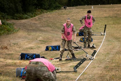 Four people in camouflage uniforms with pink vests work on metal equipment laid out on grass. The setting appears to be outdoors with trees in the background. The individuals are focused on their tasks.