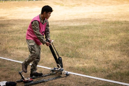 A person in camouflage clothing and a pink vest operates a small wheeled device on a grassy field, pressing down on it near a white line.